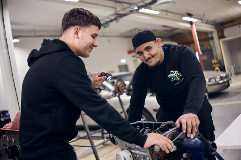 Elever står i en verkstad med bilar. En elev ser in i kameran och ler.