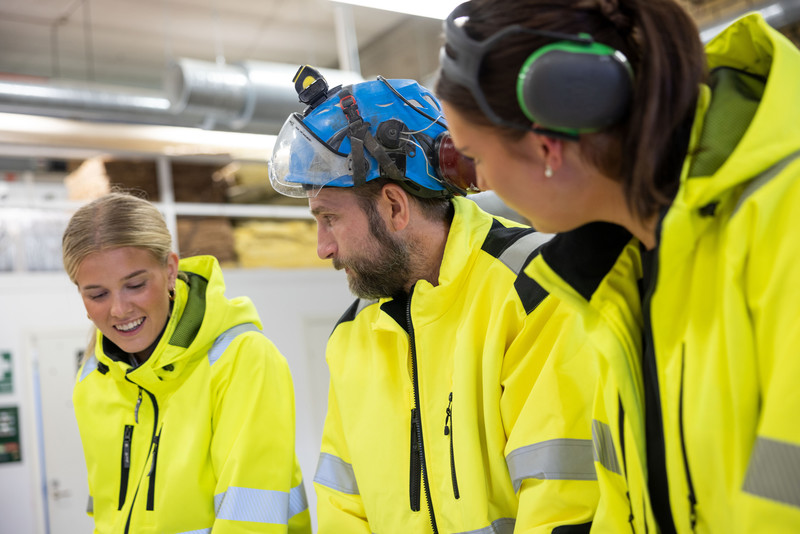 Lärare och elever i byggnadsutrustning i verkstadsmiljö.