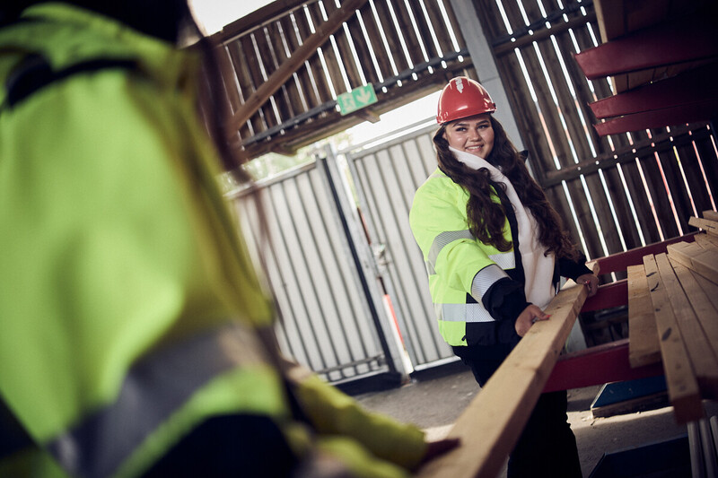 Elev står med träplanka i en verkstad och ler mot en annan elev.