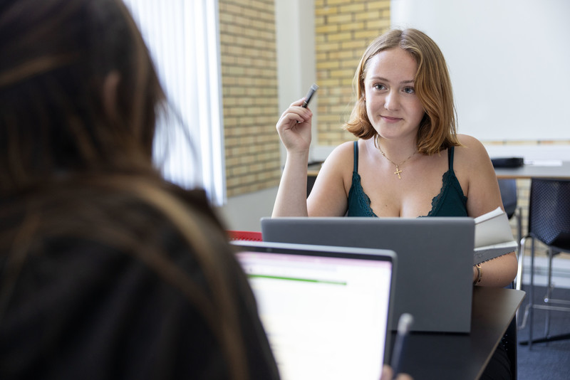 En kvinnlig elev sitter vid ett bord med laptop och penna och pratar med en annan person i en studiemiljö.
