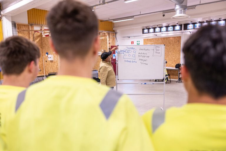 Tre elever kollar på lärare som håller i undervisning samtidigt som de tittar på lärarens anteckningar på whiteboard där läraren illustrerar olika begrepp.