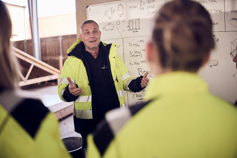 Lärare i gul skyddsutrustning står vid whiteboard och håller i undervisning för elever.