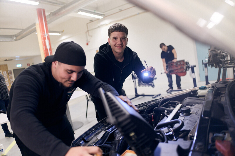 Två elever på personbils inriktningen inspekterar motorhuv på bil och lyser med ficklampa.