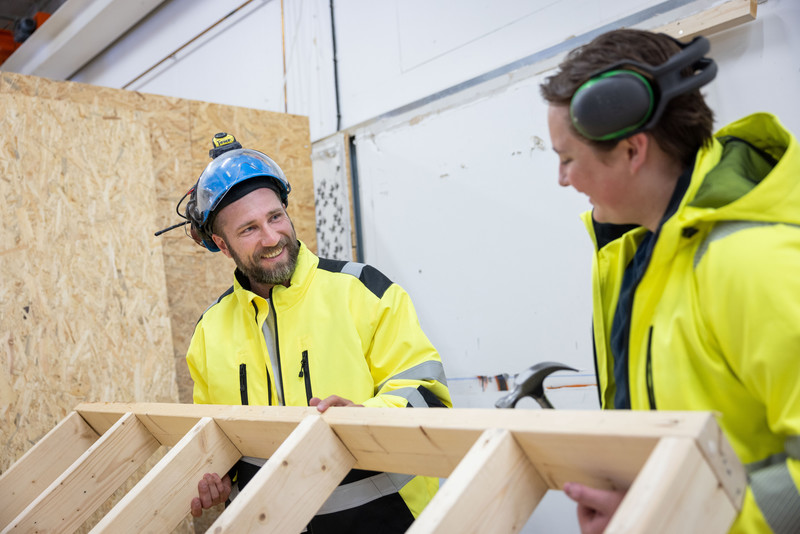 Instruktör och elev är i en verkstadsmiljö och håller i flera plankor.