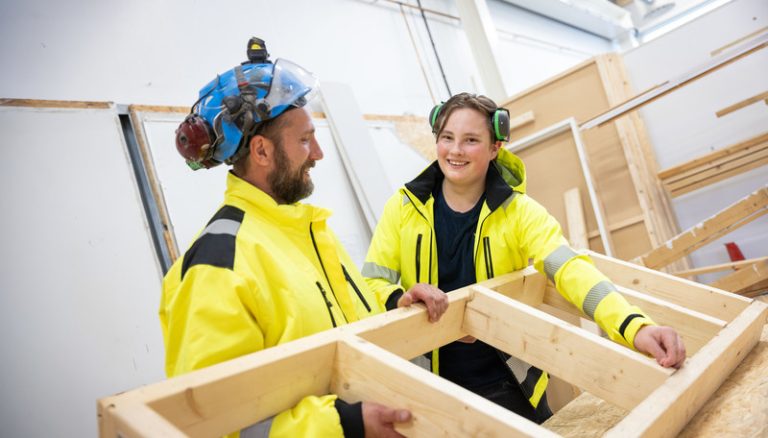 En manlig skolelev och lärare håller i flera stora brädor som de bygger på. De har på sig byggnadsutrustning, hörselkåpor och hjälm.