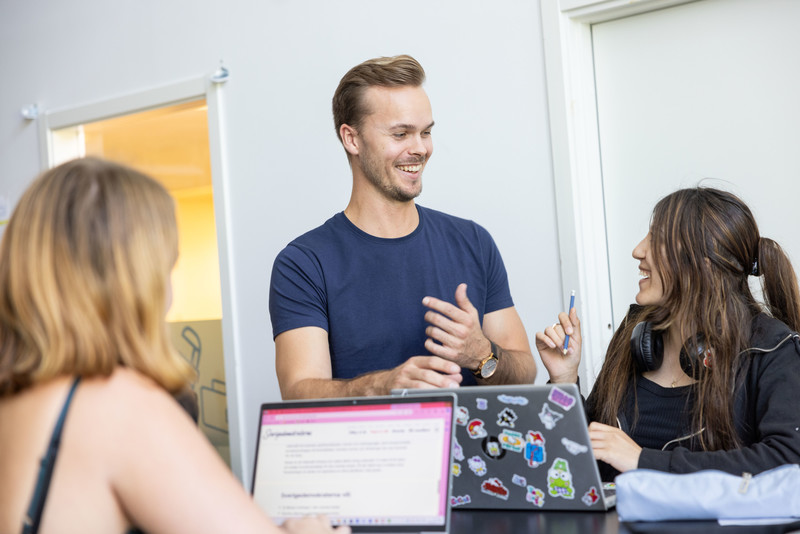 Lärare står och pratar med två elever som sitter ner vid sina datorer.
