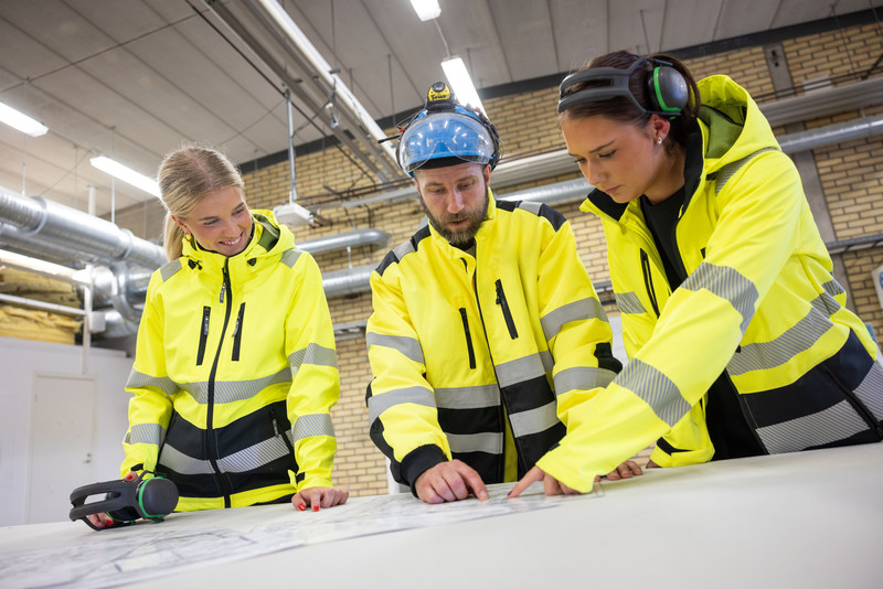 Tre personer i varselkläder tittar på ritningar inomhus. En instruktör visar och två elever lyssnar och pekar.