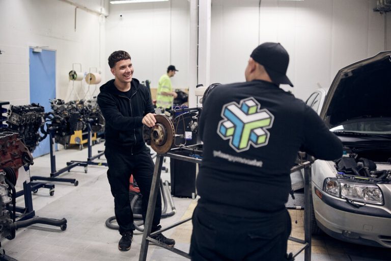 Två manliga elever i verkstadsmiljö står vid flera bildelar.