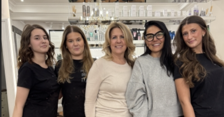 Fem personer står tillsammans och ler i en frisörsalong framför en disk och hyllor med hårprodukter. Bilden är inzoomad.