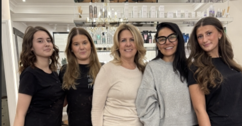 Fem personer står tillsammans och ler i en frisörsalong framför en disk och hyllor med hårprodukter. Bilden är inzoomad.