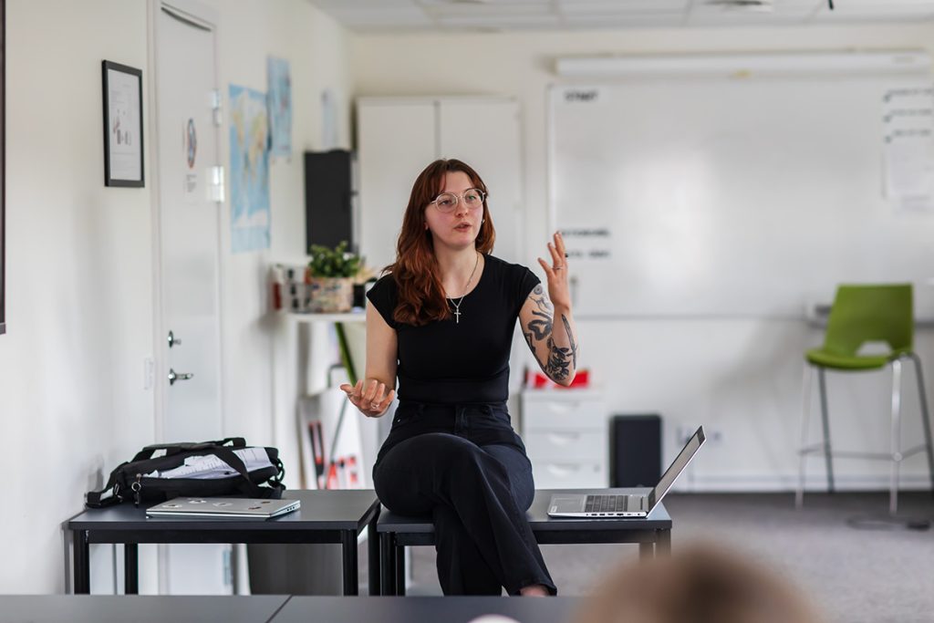 Person sitter längst fram i ett klassrum och håller en presentation. En whiteboard och bord syns i bakgrunden.