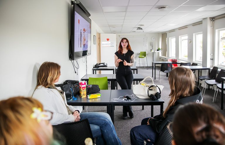 Person presenterar för en mindre grupp deltagare i ett klassrum. En skärm med presentationsmaterial visas på väggen.