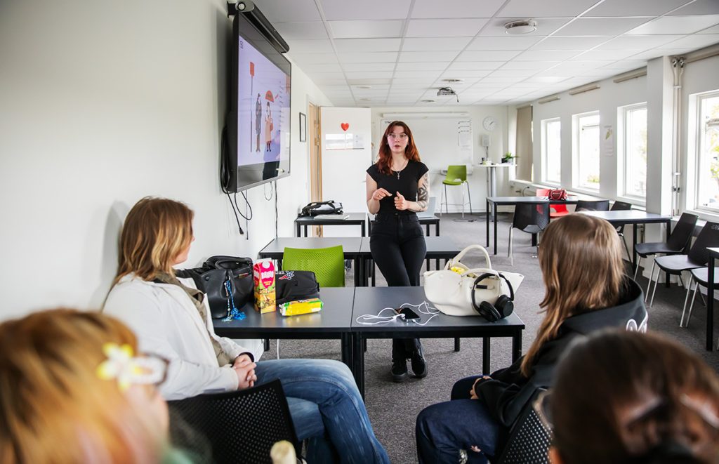 Person presenterar för en mindre grupp deltagare i ett klassrum. En skärm med presentationsmaterial visas på väggen.